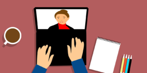 Hands lay on a laptop keyboard while a person is on the screen and a notepad is next to the laptop.
