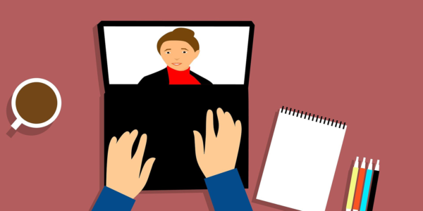Hands lay on a laptop keyboard while a person is on the screen and a notepad is next to the laptop.