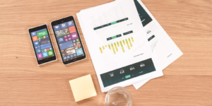 A desk with 2 cell phones, a stack of papers, and a glass of water.