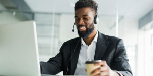 A man sitting at a desk in front of a computer with a headset on and holding a cup of coffee