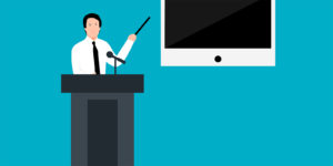 A person in a shirt and tie standing behind a podium pointing at a large screen.