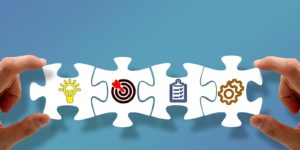 4 white puzzle pieces put together and being held between 2 hands. Each piece has a different symbol on it, including a target, gears, a checklist, and a lightbulb.