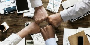 4 people fist bumping over a table containing a group of papers, computers, and phones.