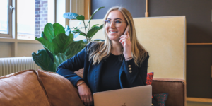 A woman is smiling while sitting on the couch wearing a blue jacket with a laptop in her lap and her ear to her phone.