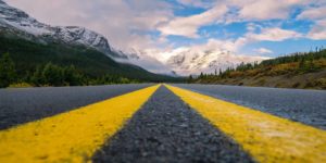 Two yellow lines come together to point down the road towards a mountain and cloudy skies