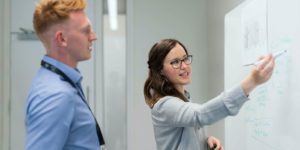 A woman shows a man the plan that she has while pointing to a whiteboard