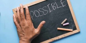Chalkboard with the word possible written on it with a hand covering a portion of the board and chalk in the corner.
