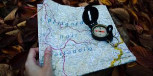 A person holds a folded map with a compass on top of it laying on a bed of leaves.