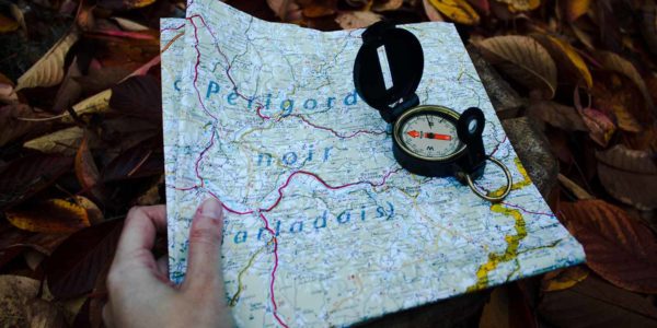 A person holds a folded map with a compass on top of it laying on a bed of leaves.