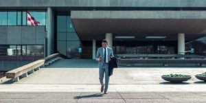 A man in a suit looks at his phone with a building behind him.