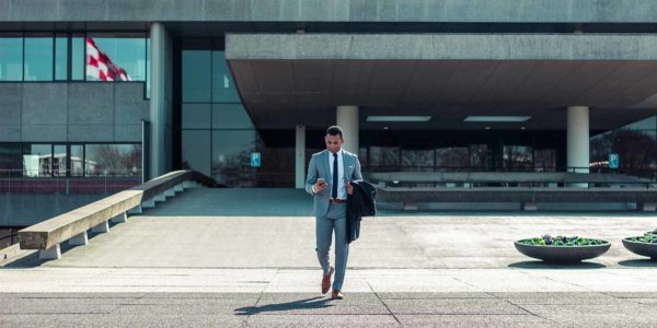 A man in a suit looks at his phone with a building behind him.