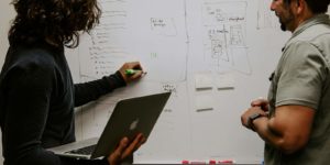 Two people stand in front of a busy whiteboard one is watching while the other writes and holds a laptop.