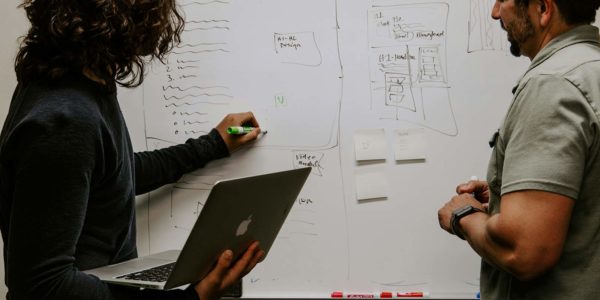 Two people stand in front of a busy whiteboard one is watching while the other writes and holds a laptop.