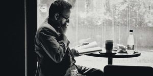 A man sits at a table with a book and pen in hand, thinking.