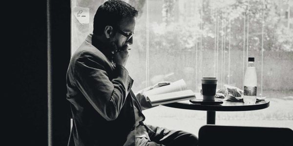 A man sits at a table with a book and pen in hand, thinking.