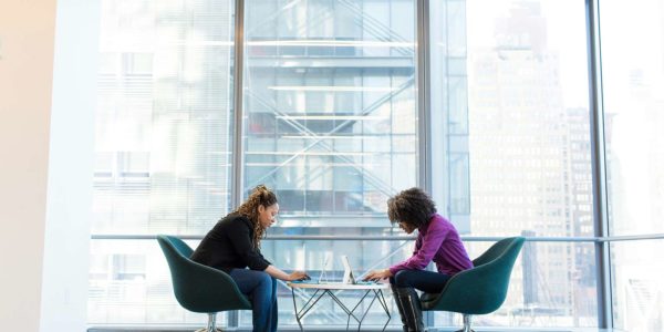 Two women sit across a table from one another and look at their laptops.