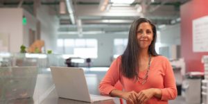 A woman stands at desk and looks at the camera with a smile.