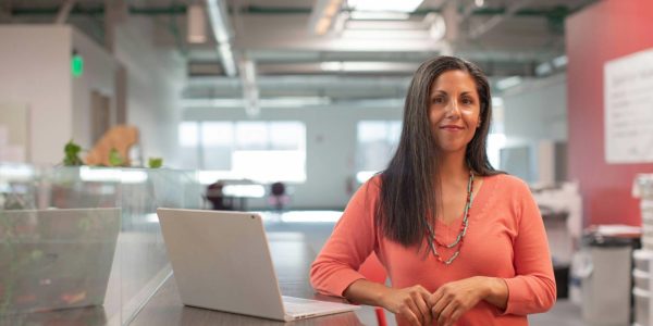 A woman stands at desk and looks at the camera with a smile.