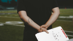 A football coach holds a whiteboard behind his back on the football field.