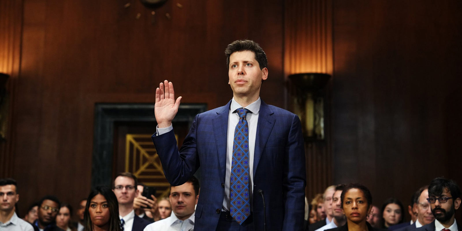 Man in blue suit stands with his right hand in the air in a court room. 