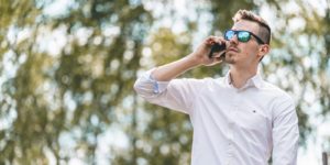 Man in white shirt and blue sunglasses stands outside on the phone.