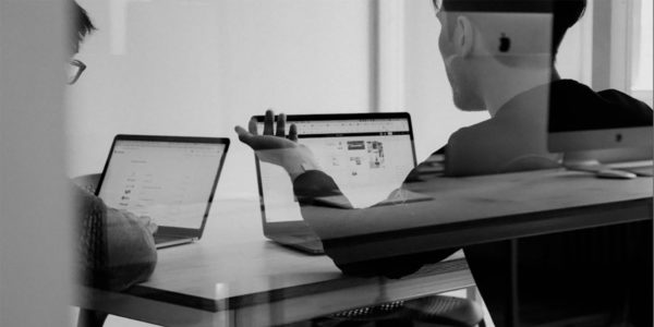 Black and white photo of two people collaborating at a table with laptops in a modern office setting