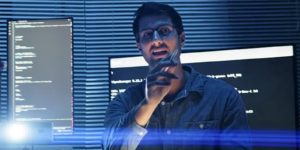 Person in glasses coding at night with blue lighting and multiple monitors displaying programming code