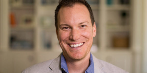 Portrait of Shawn Achor, bestselling author and positive psychology researcher, smiling in a light gray blazer