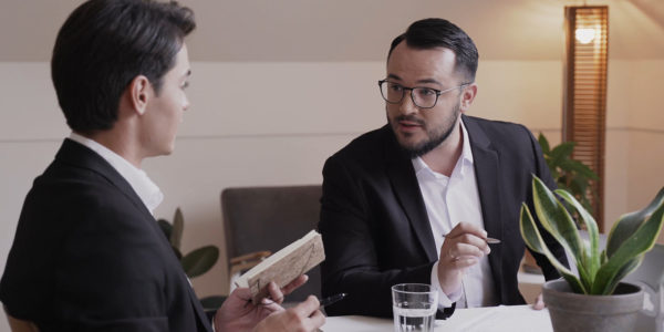 Professional_Business_Meeting_Two_People_Discussing_Strategy_Interview_Or_Collaboration_In_Office Two people in suits having a professional discussion at a desk with notes, water glass, and indoor plants