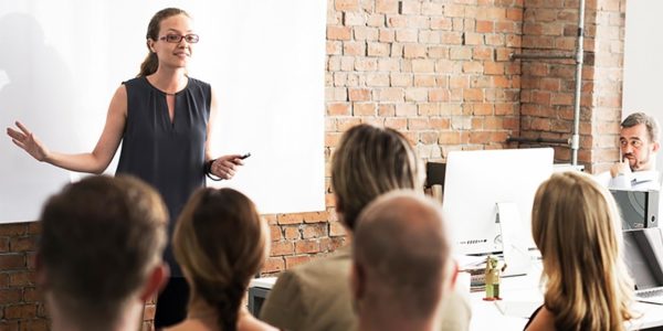 Professional woman giving a presentation to colleagues seated in a modern office with brick walls and computers visible