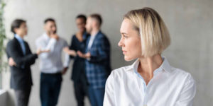 Professional woman standing apart and looking away while four colleagues talk together in the background, symbolizing isolation