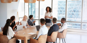 Professional woman standing and speaking to colleagues during a business meeting in a modern office with large windows and city view